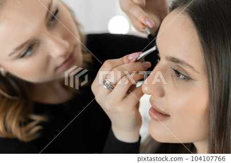 The photo captures an experienced makeup artist perfecting the client's eyebrows. Every detail counts and the master focuses on creating perfect symmetry and close-up lines in a beauty salon. The photo captures an experienced makeup artist perfecting the client's eyebrows. Every detail counts and the master focuses on creating perfect symmetry and close-up lines in a beauty salon. 104107766