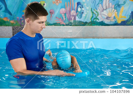 Trainer teaching little boy how to swim in indoor pool with pool floating board 104107896