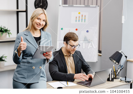 Cheerful young corporate lawyers in formal wear editing text of new contract using laptop sitting at table with laptop in modern office. Woman sits next to a colleague and holds a tablet in her hands 104108455