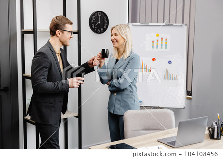 Couple of cheerful colleagues talking while standing at the flipchart in office with cups of coffee 104108456