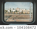 Train window view with old railway, clear blue sky, white and red brick buildings, yellow and red freight wagons, metal fence, city wires and industrial area. Pale non-contrast color correction. 104108472