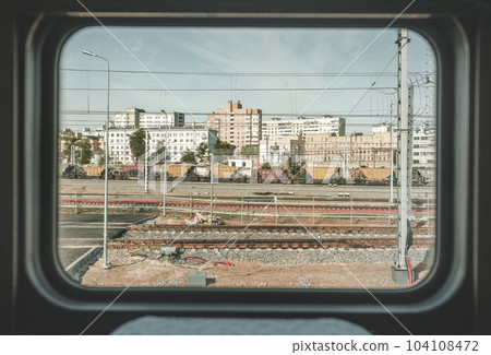 Train window view with old railway, clear blue sky, white and red brick buildings, yellow and red freight wagons, metal fence, city wires and industrial area. Pale non-contrast color correction. 104108472