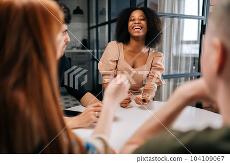 Laughing black female playing fun cards games for time with diverse friends, enjoying pastime activity sitting at table at home. Happy multiethnic men and women playing board game in living room. 104109067