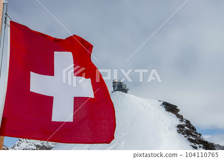 Swiss flag waving on pole with view of Jungfrau ice palace on top of snow covered mountain under cloudy sky at Switzerland, winter holiday travel concept 104110765