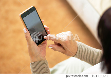 Close up of hands from middle aged woman sitting on sofa at home and using a mobile phone. 104111652