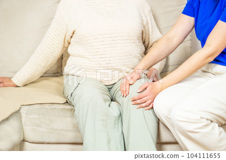 Young caregiver helping senior woman with knee pains at home. Caring nurse assisting her patient. Young caregiver helping senior woman with knee pains at home. Caring nurse assisting her patient. 104111655