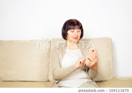 Happy relaxed older woman sitting on comfortable sofa at home and using a mobile phone. 104111670
