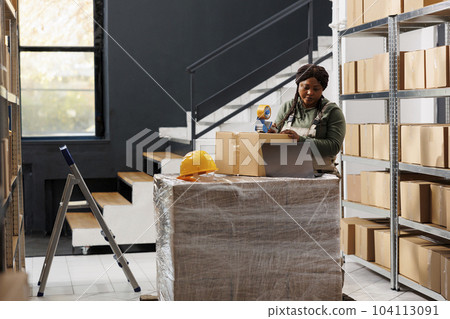 Stockroom worker using adhesive tape to pack clients orders, preparing packages for shipping in storage room. African american supervisor wearing industrial overall during goods inventory 104113091