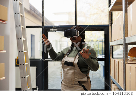 Storage room employee wearing virtual reality headset, enjoying warehouse simulation, preparing customers orders in storehouse. African american supervisor working at merchandise inventory 104113108
