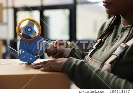 Storage room worker putting adhesive tape on carton boxes, preparing customers orders in warehouse. African american employee checking products before delivery, working in storage room. Close up Storage room worker putting adhesive tape on carton boxes, preparing customers orders in warehouse. African american employee checking products before delivery, working in storage room. Close up 104113127