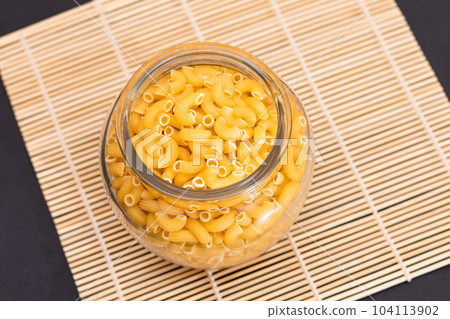 Uncooked Chifferi Rigati Pasta in Glass Jar on Bamboo Mat on Black Background 104113902
