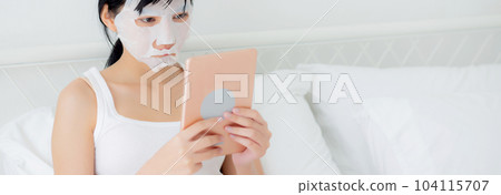 Beautiful young asian woman with sheet facial mask sitting watching tablet computer on bed. 104115707