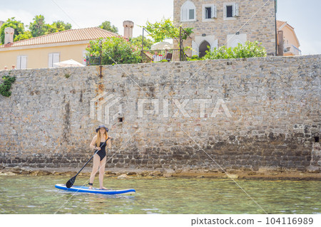 Young women Having Fun Stand Up Paddling in blue water sea in Montenegro. Against the backdrop of the Old Town of Budva. SUP 104116989