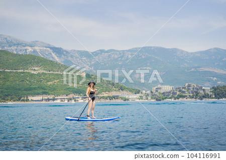 Young women Having Fun Stand Up Paddling in blue water sea in Montenegro. Against the backdrop of the Montenegrin mountains. SUP 104116991