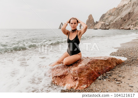 Woman travel portrait. Happy woman with long hair looking at camera and smiling. Close up portrait cute woman in black bikini posing on a red volcanic rock on the sea beach Woman travel portrait. Happy woman with long hair looking at camera and smiling. Close up portrait cute woman in black bikini posing on a red volcanic rock on the sea beach 104121617