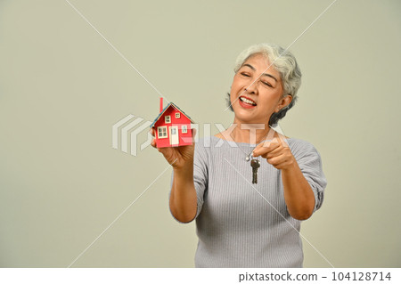 Smiling mature woman holding house model and keys isolated on beige background. Real estate, building, property and insurance Smiling mature woman holding house model and keys isolated on beige background. Real estate, building, property and insurance 104128714