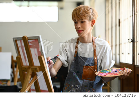 Peaceful young stylish gay man painting picture with oil paints on canvas in bright art studio. Leisure activity and art concept 104128719