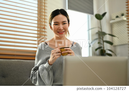 Smilie millennial woman drinking herbal tea, enjoy leisure weekend at home, browsing internet on laptop Smilie millennial woman drinking herbal tea, enjoy leisure weekend at home, browsing internet on laptop 104128727