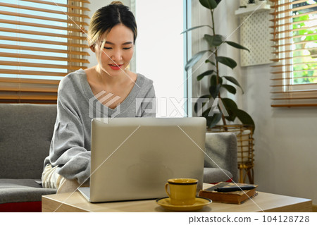 Beautiful asian woman sitting on couch, working remotely, surfing internet on laptop at home 104128728