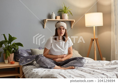 Horizontal shot of sick unhealthy sad woman wearing sleeping mask sitting on bed in cozy bedroom, having terrible strong stomachache, frowning face from pain. Horizontal shot of sick unhealthy sad woman wearing sleeping mask sitting on bed in cozy bedroom, having terrible strong stomachache, frowning face from pain. 104129894