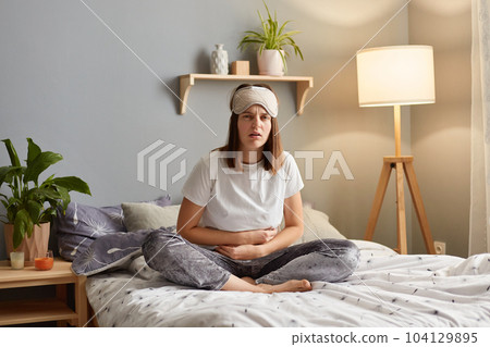 Portrait of upset Caucasian woman suffering from strong abdominal pain, touching her tummy, sitting on bed, feeling acute stomachache, has chronic disease, spasm, or hernia. Portrait of upset Caucasian woman suffering from strong abdominal pain, touching her tummy, sitting on bed, feeling acute stomachache, has chronic disease, spasm, or hernia. 104129895