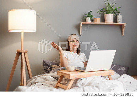 Portrait of confused uncertain woman wearing sleeping mask working on laptop from home while lying on bed, having video call with partner, online meeting, dosen't know what to do, spreads hand aside. Portrait of confused uncertain woman wearing sleeping mask working on laptop from home while lying on bed, having video call with partner, online meeting, dosen't know what to do, spreads hand aside. 104129935