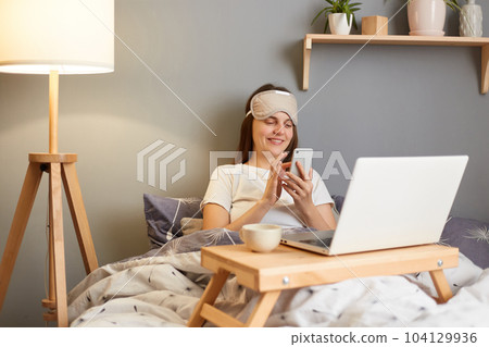 Photo of young adult brown haired woman wearing sleep mask sitting with laptop in bed at home and typing on mobile phone, writing message or scrolling online. 104129936