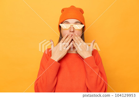 Horizontal shot of amazed shocked surprised woman wearing orange sweater, beanie hat and glasses in minecraft style standing isolated over yellow background, covering mouth with palms. 104129953