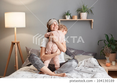 Joyful mother in sleeping mask on her forehead hugging her infant baby daughter playing in bed at, woman embracing her child, being happy to spend morning together, family playing after wake up. Joyful mother in sleeping mask on her forehead hugging her infant baby daughter playing in bed at, woman embracing her child, being happy to spend morning together, family playing after wake up. 104130189