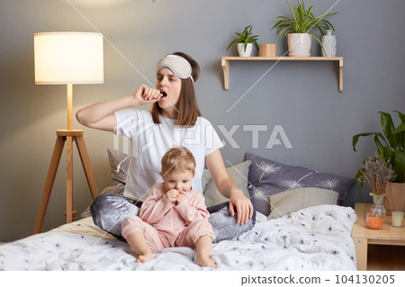 Indoor shot of sleepy exhausted woman wearing pajama and blindfold with her baby daughter in the bedroom sitting on bed, playing with toddler kid, yawning, covering mouth with palm. Indoor shot of sleepy exhausted woman wearing pajama and blindfold with her baby daughter in the bedroom sitting on bed, playing with toddler kid, yawning, covering mouth with palm. 104130205