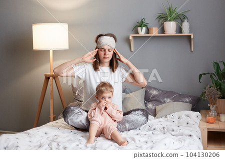 Portrait of sick tired Caucasian mother and her ingfant daughter sitting together on bed, woman having headache, massaging her temples, feels pain in head after sleepless night. Portrait of sick tired Caucasian mother and her ingfant daughter sitting together on bed, woman having headache, massaging her temples, feels pain in head after sleepless night. 104130206