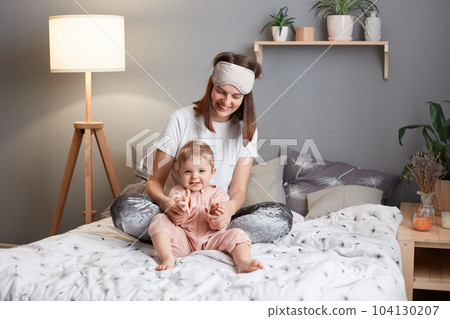 Photo of joyful cheerful mother and child laughing and playing together while sitting on bed, woman wearing pajama and sleeping mask having fun with her infant kid. Photo of joyful cheerful mother and child laughing and playing together while sitting on bed, woman wearing pajama and sleeping mask having fun with her infant kid. 104130207