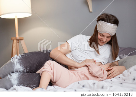Indoor shot of young beautiful mother, breastfeeding her newborn baby girl in the morning, woman in blindfold on her forehead lying in bed and nursing her infant daughter. 104130213