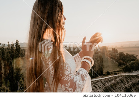 Happy woman in white boho dress on sunset in mountains. Romantic woman with long hair standing with her back on the sunset in nature in summer with open hands. Silhouette. Nature. Sunset. 104130369