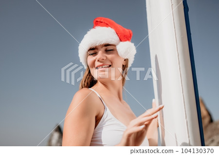 Woman sea sup. Close up portrait of happy young caucasian woman with long hair in Santa hat looking at camera and smiling. Cute woman portrait in a white bikini posing on sup board in the sea 104130370