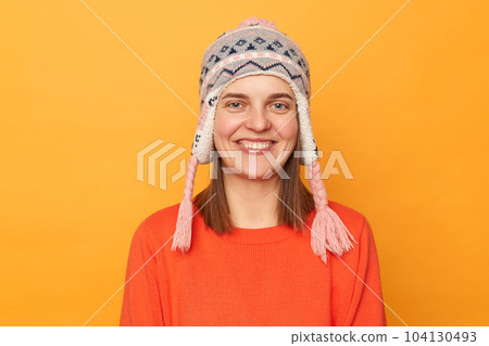 Portrait of smiling beautiful woman wearing orange jumper and knitted earflap hat standing isolated over yellow background, looking at camera with toothy smile, being in good mood. 104130493