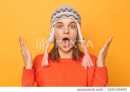 Portrait of astonished young adult Caucasian woman wearing funny earflap hat and orange casual jumper standing isolated over yellow background, raised her arms, being shocked. 104130495