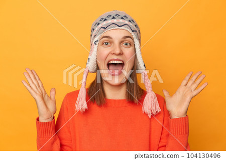 Indoor shot of excited positive happy woman wearing orange jumper and knitted earflap hat standing isolated over yellow background, raised her arms, screaming with happiness. 104130496