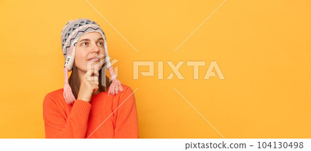 Indoor shot of pensive woman wearing orange sweater and knitted earflap hat standing isolated over yellow background, looking away at advertisement area, holding chin, having thoughtful expression. 104130498