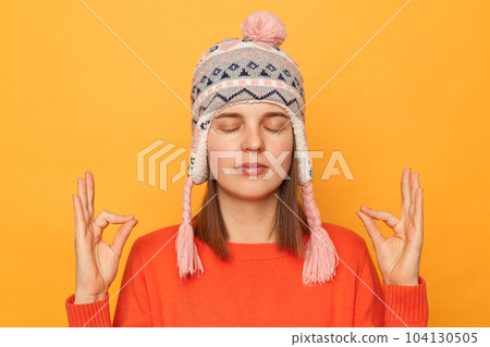 Portrait of peaceful calm woman wearing orange jumper and knitted earflap hat, standing with closed eyes, doing yoga, meditating, standing isolated over yellow background. 104130505