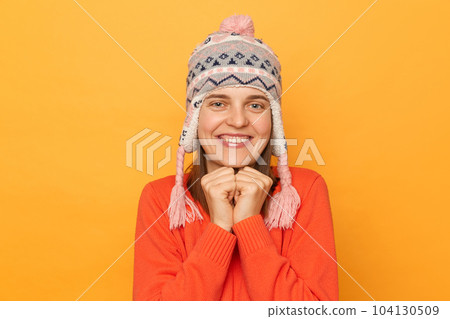 Portrait of cute delighted Caucasian woman wearing funny earflap hat and orange casual jumper, looking at camera, keeps fists under chin, standing isolated over yellow background. 104130509