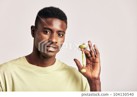 African guy in a t-shirt with jade de-puffing face roller. Black man in studio with beauty, health and natural skin procedure. Wellness, cosmetics and skin care for men. Close up portrait. 104130540