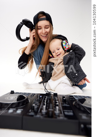 A young pretty barefoot mother in a black top, blue jeans and a black baseball cap and her baby in a black motorcycle jacket are sitting on the floor. They play with DJ headphones and mixing board. 104130599