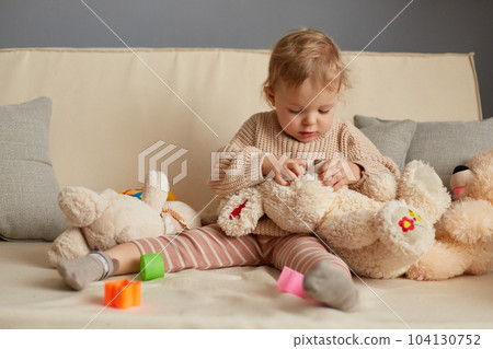Image of attentive concentrated baby girl in beige sweater sitting on cough with a plush big rabbit, trying to find nose, looking at soft toy with concentrated expression. 104130752