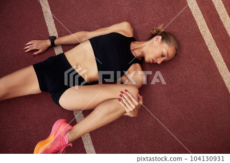 A young athlete girl is warming up before training at the stadium. A slender blonde woman in a black tracksuit does exercises to stretch her muscles while lying on a treadmill. Sports and recreation. A young athlete girl is warming up before training at the stadium. A slender blonde woman in a black tracksuit does exercises to stretch her muscles while lying on a treadmill. Sports and recreation. 104130931