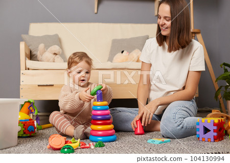 Indoor shot of kid playing with pyramid toy together with mother, happy little girl collects a toy pyramid of rings on floor in a kindergarten, development of fine motor skills in children lifestyle. Indoor shot of kid playing with pyramid toy together with mother, happy little girl collects a toy pyramid of rings on floor in a kindergarten, development of fine motor skills in children lifestyle. 104130994