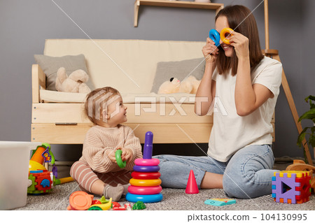 Indoor shot of funny mother wearing white shirt and jeans playing with her little daughter at home while sitting on floor, mommy covering her eyes with rings of pyramid. Indoor shot of funny mother wearing white shirt and jeans playing with her little daughter at home while sitting on floor, mommy covering her eyes with rings of pyramid. 104130995