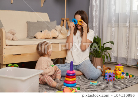 Activities with children at home, early childhood development. Young adult woman sitting on floor in living room, playing with her baby child with pyramid, covering her eyes with rings, having fun. Activities with children at home, early childhood development. Young adult woman sitting on floor in living room, playing with her baby child with pyramid, covering her eyes with rings, having fun. 104130996
