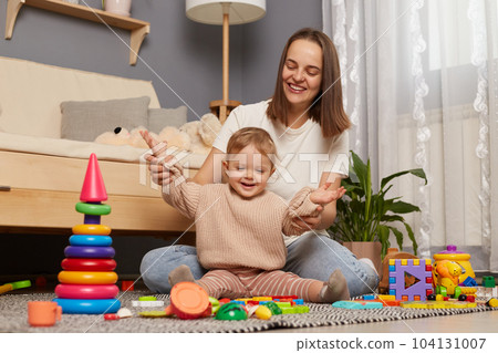 Indoor shot of happy mother with her infant daughter playing together, rejoicing their results with early development, sittng on floor in living room. Motherhood and childhood. 104131007