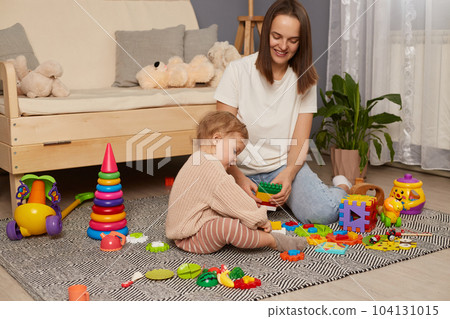 Mom and child build out of colorful plastic blocks, smiling woman and her daughter in playroom making brick constructions, early development, preschooler education. Mom and child build out of colorful plastic blocks, smiling woman and her daughter in playroom making brick constructions, early development, preschooler education. 104131015